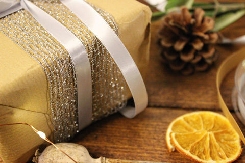 close up of a gift wrapped in brown paper with silver ribbon and lurex mesh braid around it. It is surrounded by festive odds and ends like pine cones, ribbon, pine sprigs and tiny fairy lights.