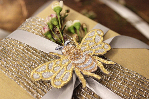 A close up of a gift wrapped in brown paper with a silver lurex net braid and silver double sided ribbon around it. It is topped with a small embroidered silver and gold bee motif and a sprig of waxflower