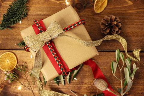 A gift is wrapped in brown paper with red ribbon and a red, green and gold chunky cord around it. It is topped with a sparkly gold bow. It is surrounded by festive odds and ends like pine cones, ribbon, pine sprigs and tiny fairy lights.
