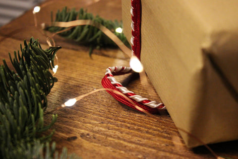 A close up of the corner of a gift wrapped in brown paper, focussing on a red and white candy striped trimming. There also tiny fairy lights and sprigs of pine in shot.