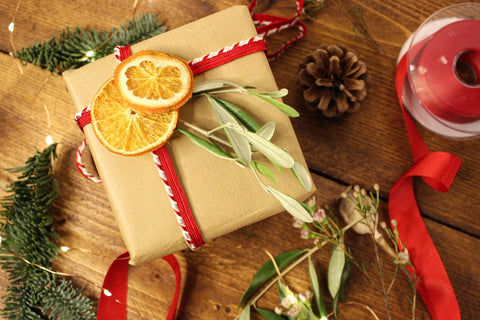 A gift is wrapped in brown paper with a red and white candy striped braid wrapped around it with dried oranges and an olive branch decorating it. It is surrounded by festive odds and ends like pine cones, ribbon, pine sprigs and tiny fairy lights.7482 insertion 6503 ribbon