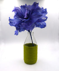 A milk bottle that has been wrapped from the shoulders down in green twine sits on a white backdrop. It has a royal blue feather flower inside it, to show it being used as a vase