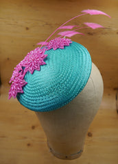 A turquoise blue circular straw hat base sits jauntily on a mannequin head. On the base, there are several hot pink beaded motifs in a starburst design, and 5 arrowhead design ostrich feather motifs, also in hot pink