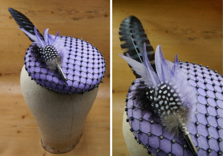 A purple button felt hat base is covered in black and diamante veiling and topped with a fancy feather motif in black and natural feather tones