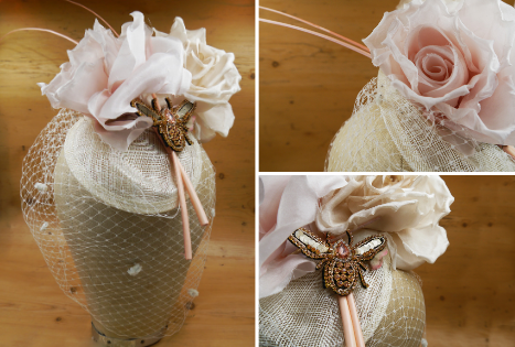 A millinery creation of a blush pink sinamay hat base, topped with large pink faux roses and pale spotted veiling. In between the roses are 2 pale pink ostrich quills and a diamante bee motif, coloured rose gold