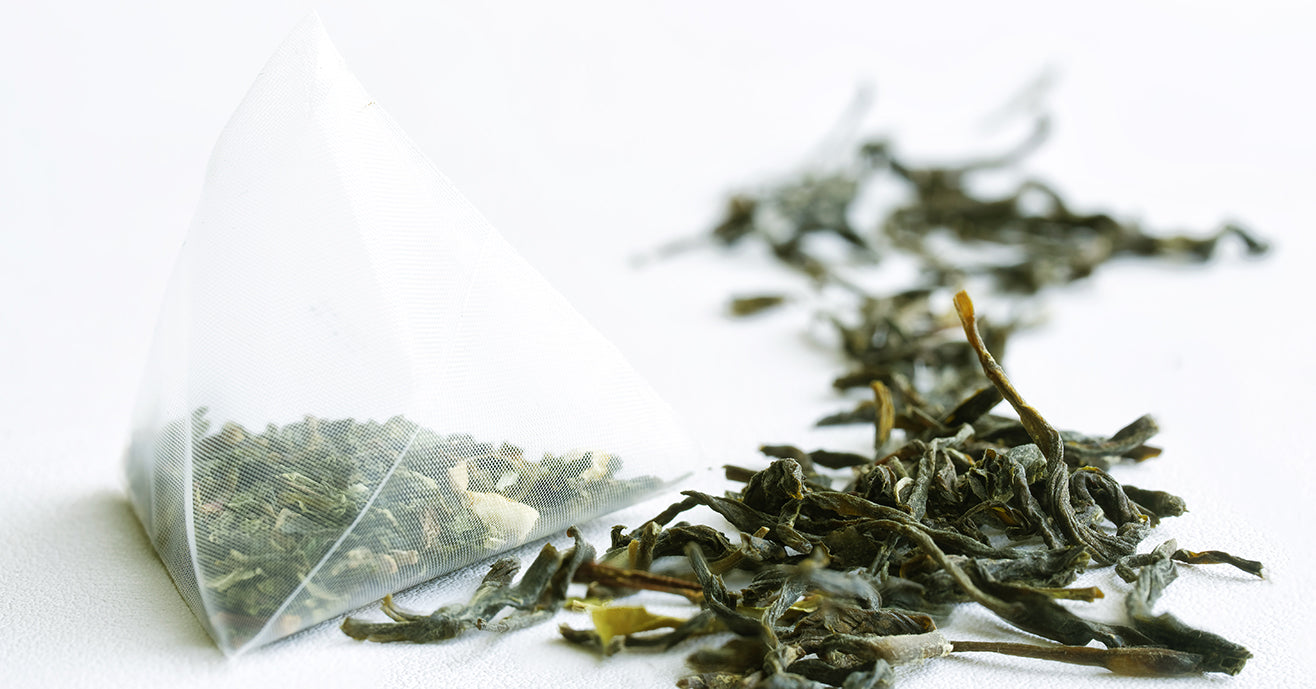 A pyramid tea bag filled with green tea, next to a mound of loose-leaf green tea.