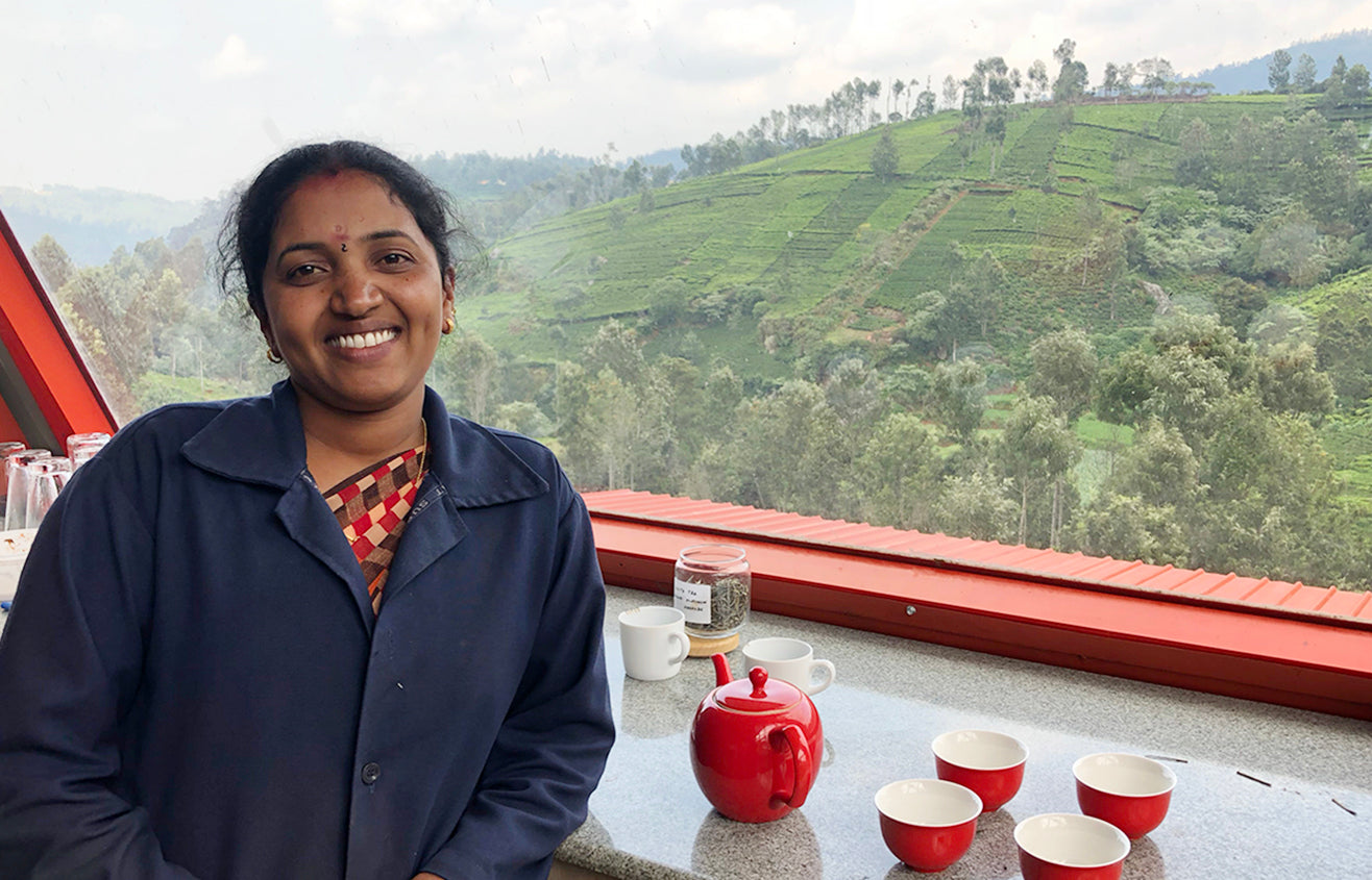 Female tea factory worker in her navy uniform leaning against a tea table, looking out into the lush tea fields, with a red tea set next to her.
