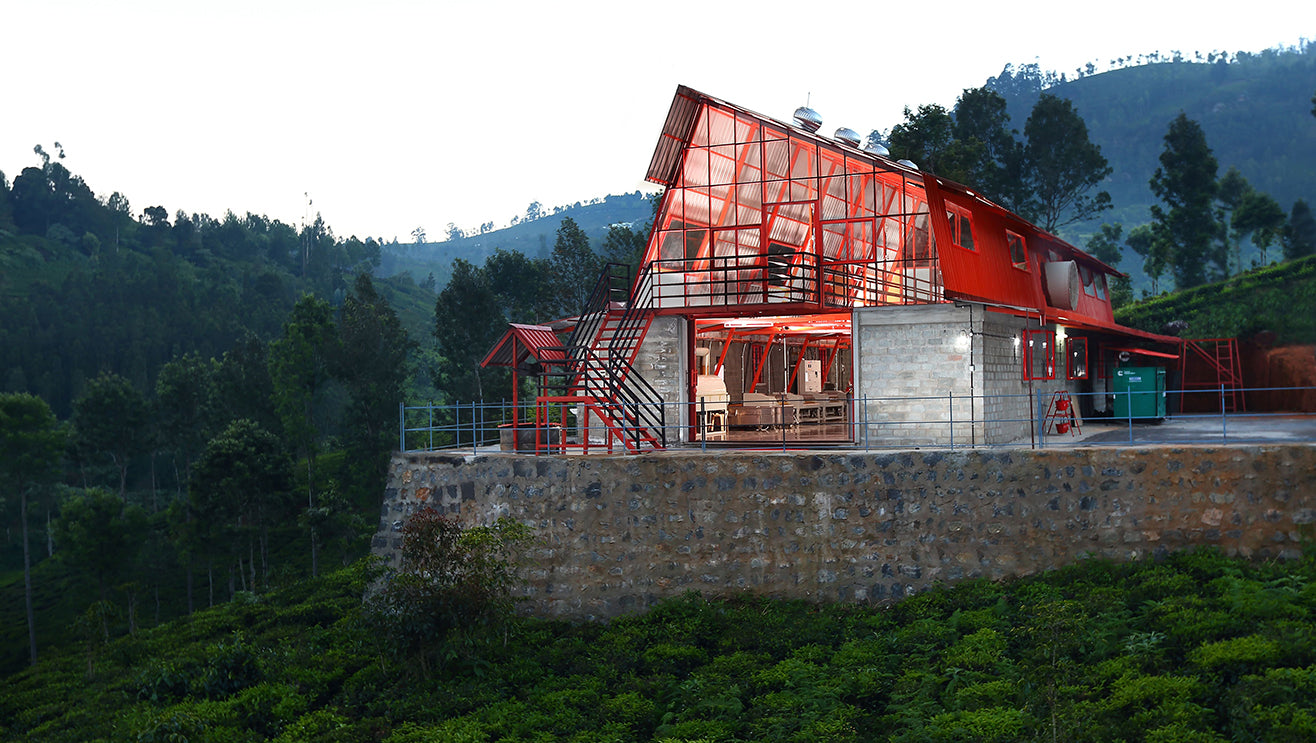 The red and modern Tea Studio Factory building in the Nilgiris at dusk, perched on top of tea fields, with angled roof and light coming from the inside.