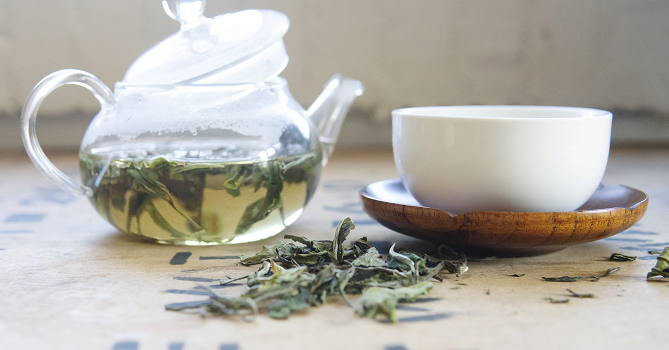 A glass teapot of white tea, next to a pile of dried white tea leaves and a white cup.