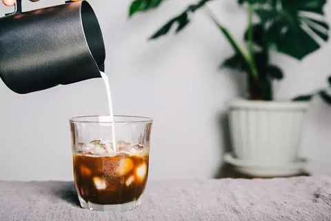 Creamer being poured into iced coffee on counter with plants in background