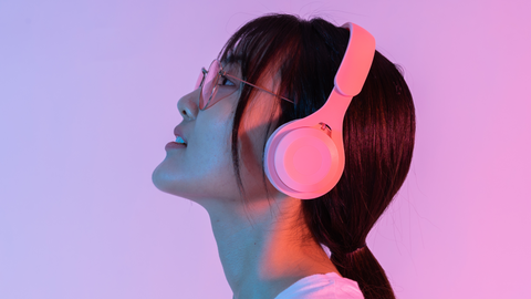 Asian femme's left side profile. they're wearing pink over the ear noise cancelling headphones. the frame is gradient light purple to pink from left to right.