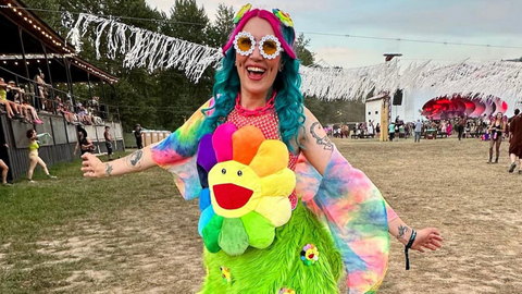 Girl with Teal and pink hair, at Bass Coast music festival, smiling big wearing a colourful flower stuffy as a top, with fun circular sunglasses made out of daisies