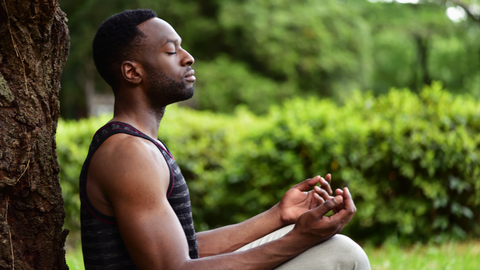 a Black man is sitting cross legged while leaned up against a tree meditating with his palms face up.