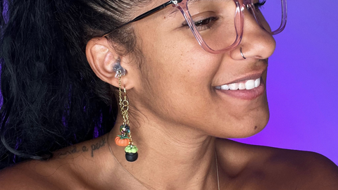 Black woman facing the right of the screen smiling wearing a pair of Sauce Strap Spooky SZN High fidelity ear plug earrings in gold. there are hand crafted pumpkin and bubbling cauldron charms dangling. the plug is in her ear. there is a purple background.