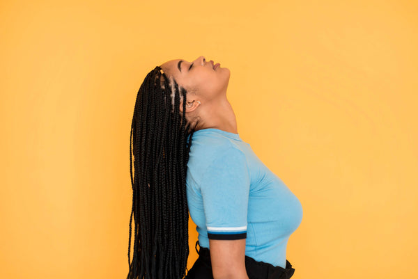 Black woman with blue shirt long braided hair standing behind yellow wall