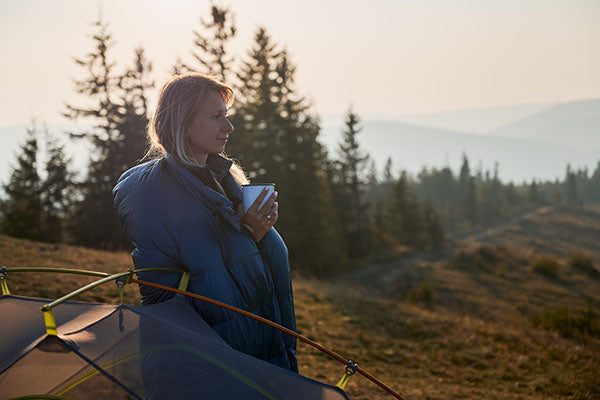 A woman wearing a sleeping bag in the morning of a camping trip.