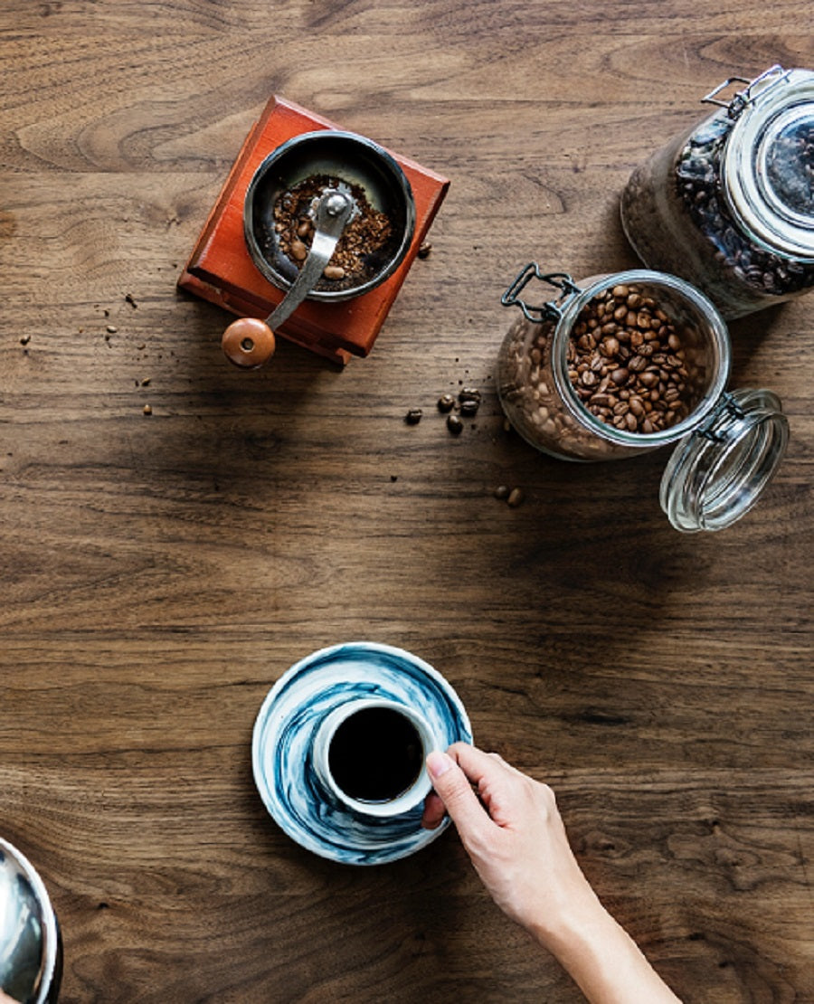 Looking down at a table with coffee beans, a hand coffee grinder, and a cup of coffee.
