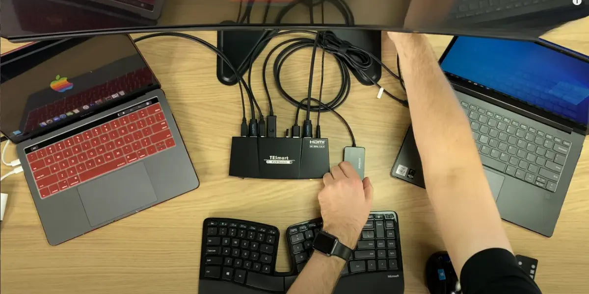 Top-down view of 2-computer KVM switch wiring setup showing HDMI and USB cables connected to a TESmart KVM, MacBook, and Windows laptop.