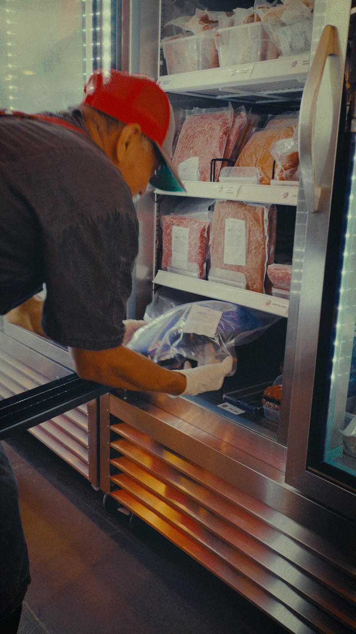 Butcher in red cap organizing packaged meat in a display fridge.