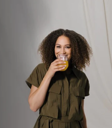 A smiling, mixed race woman with long curly hair drinking an orange drink.