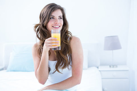 Caucasian woman drinking a glass of orange juice while sitting on a bed.