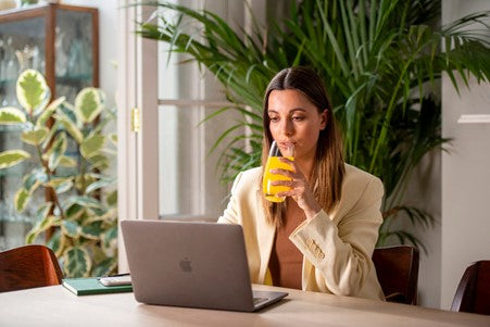 A woman sitting at a table on her laptop drinking a glass of Zest Active supplement drink