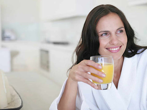 A middle-aged woman with clear, smooth skin, drinking a glass of orange juice in her dressing gown.