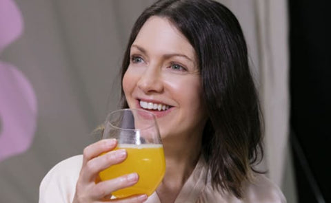 a beautiful youthful woman is looking over her glass of orange-coloured collagen drink.