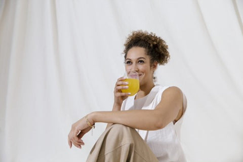 Mixed race woman sitting on the wearing neutral-coloured clothing. She drinks an orange-coloured drink with her other arm resting on her raised knee.