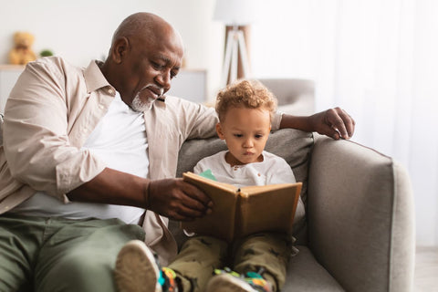 An older man sitting on the sofa, reading his grandson a book.