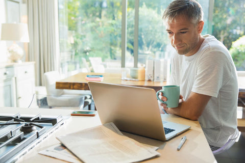 A middle-aged man sitting at a kitchen counter working at his laptop while drinking a cup of coffee.