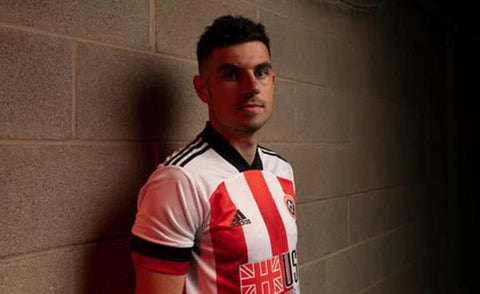 A male Sheffield United football player, leaning against a cinderblock wall in his kit.