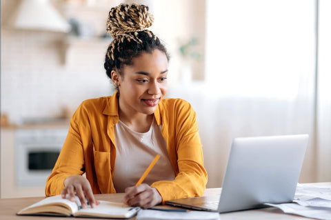 Woman wearing an orange shirt and braids is looking at her computer as she takes notes