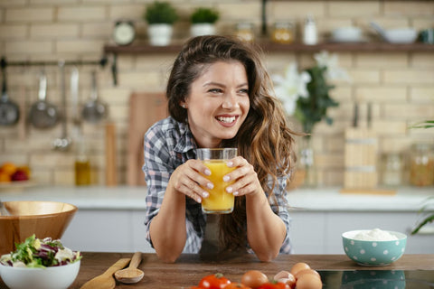 woman with long healthy hair is leaning over a kitchen counter drinking a glass of orange juice