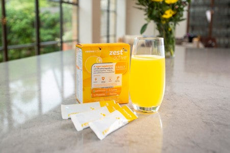 Three sachets of Zest Active are laid out on the counter in front a glass of orange drink and the supplement’s outer packaging. 