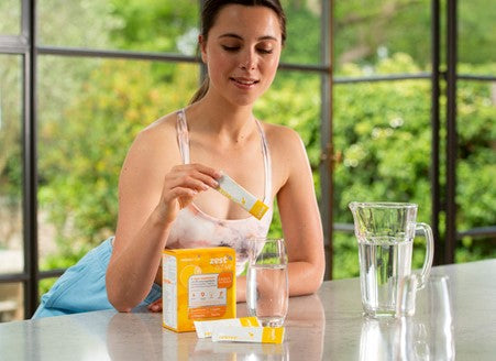 A woman in activewear, leaning onto a countertop, pouring a sachet of Zest Active into a glass of water. The box of Zest Active is sitting on the counter too.