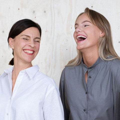 a close up of two ladies standing next to each other laughing. one has dark hair and it is tied up and the other has blonde hair and it is down