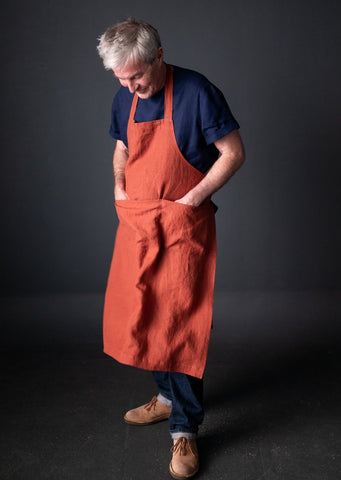 a grey haired man standing on a black background wearing a rust coloured apron the apron is long and takes up most of his body. showing from his calves down and a blue tshirt