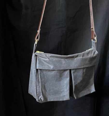a picture of a bag hanging in front of a black back ground. the bag is grey oilskin that has a rustic old patina to it