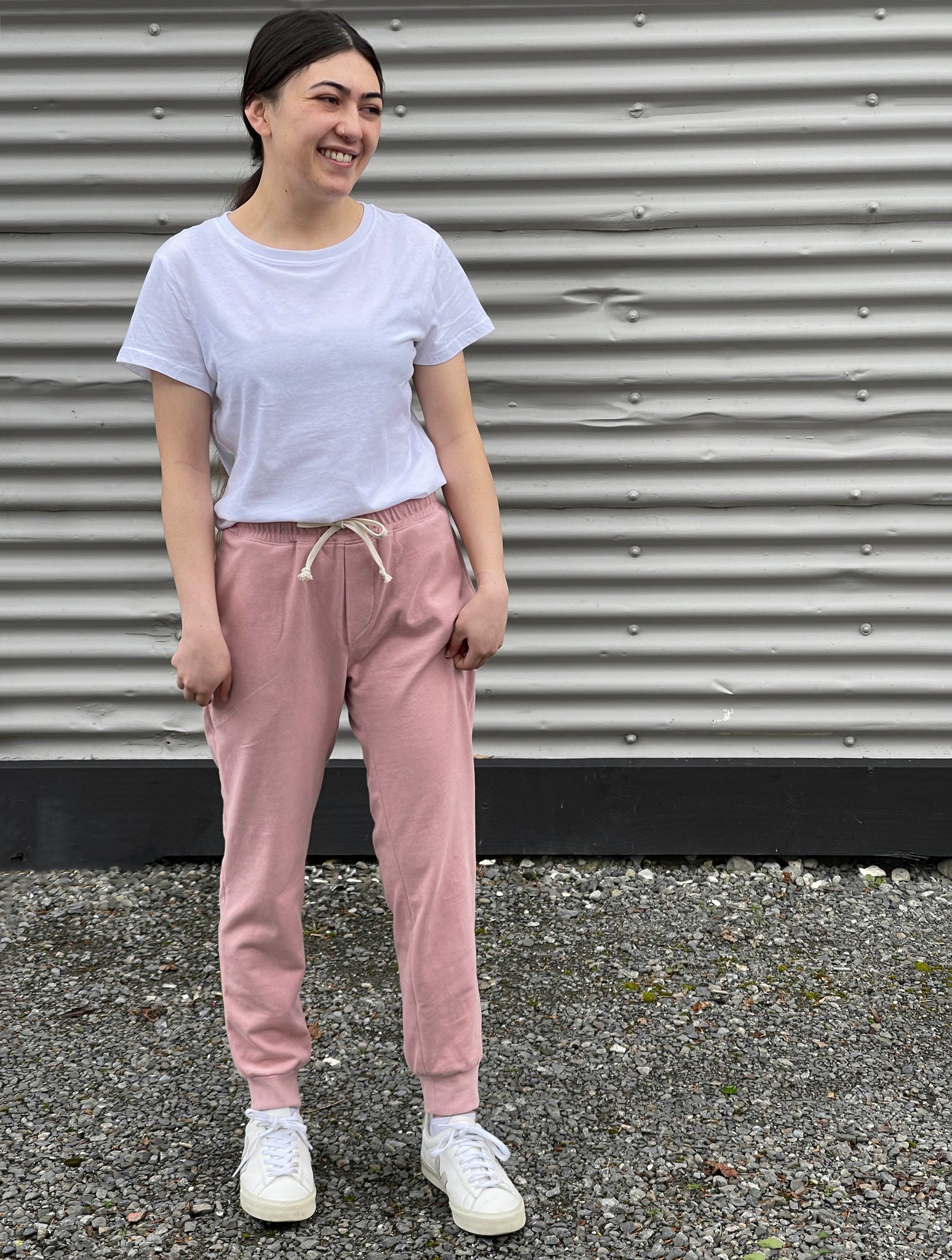 a girl is standing against a grey corrugated wearing pink jogging bottoms and a white tshirt and white trainers 