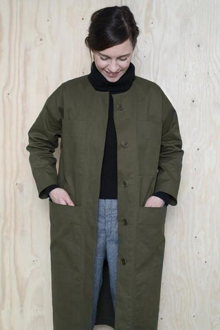 a white lady standing against a plywood background wearing a olive green long jacket with a black roll neck jumper and blue jeans underneath