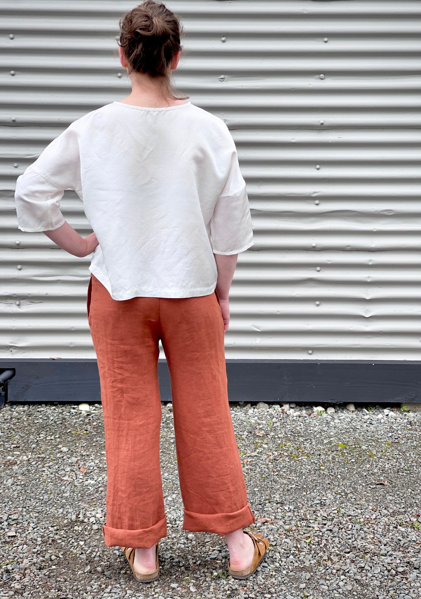 the same woman wearing the wide leg trousers and the loose white top but showing her from the back. The background is a corrugated iron wall  