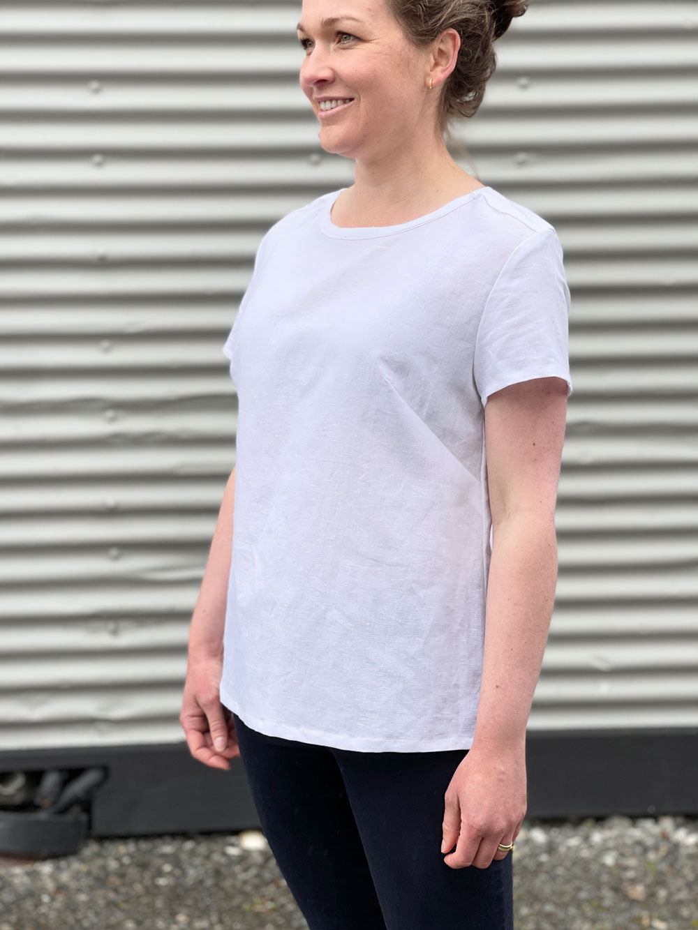 a white woman wearing a white t-shirt and black jeans with a side on shot against a corrugated iron background 