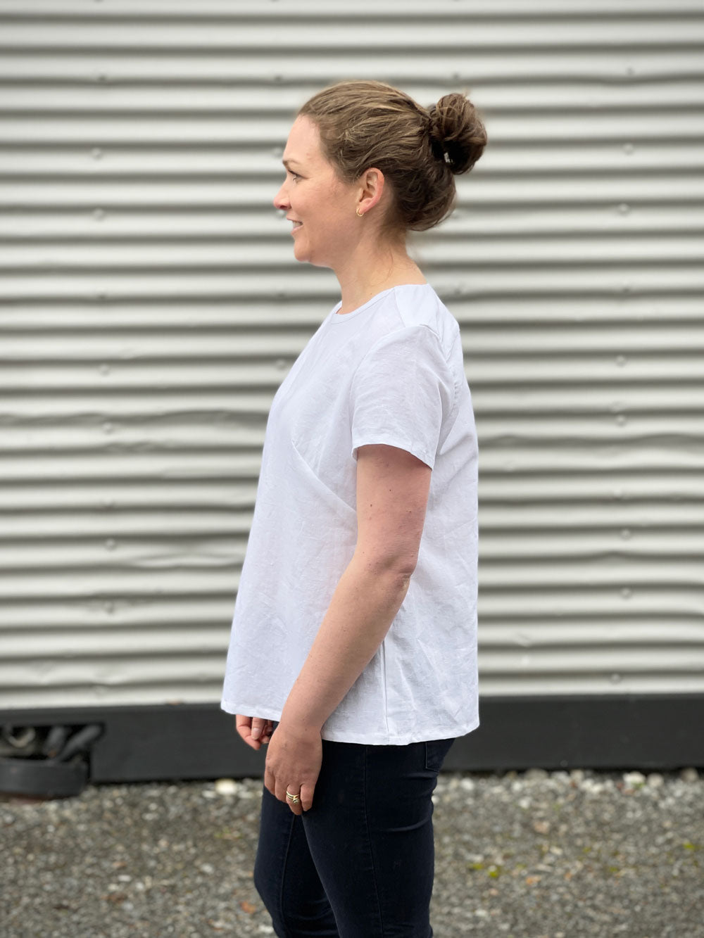 a white woman wearing a white t-shirt and black jeans with a side on shot against a corrugated iron background 