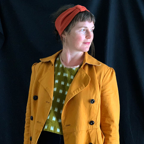 an image of a white woman wearing a dark yellow jacket and a green and white check top. she is looking to the bottom right of the screen and is wearing a rust coloured headband which she has made.