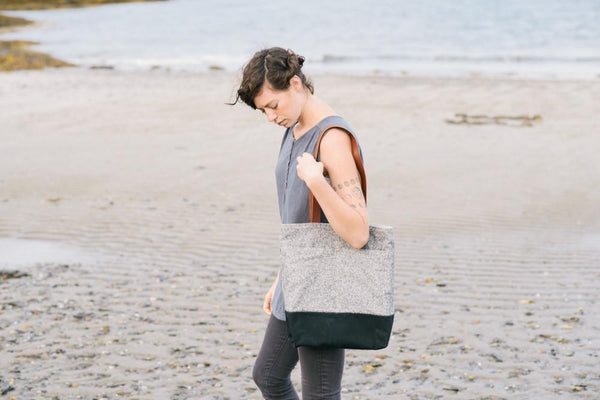 a white woman walking on a deserted beaching with a wool and wax tote bag prominently displayed, hanging over her shoulder. 