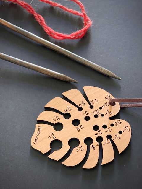 a wooden needle gauge in the shape of a monstera leaf on a black background with two needles and some red wool in the background