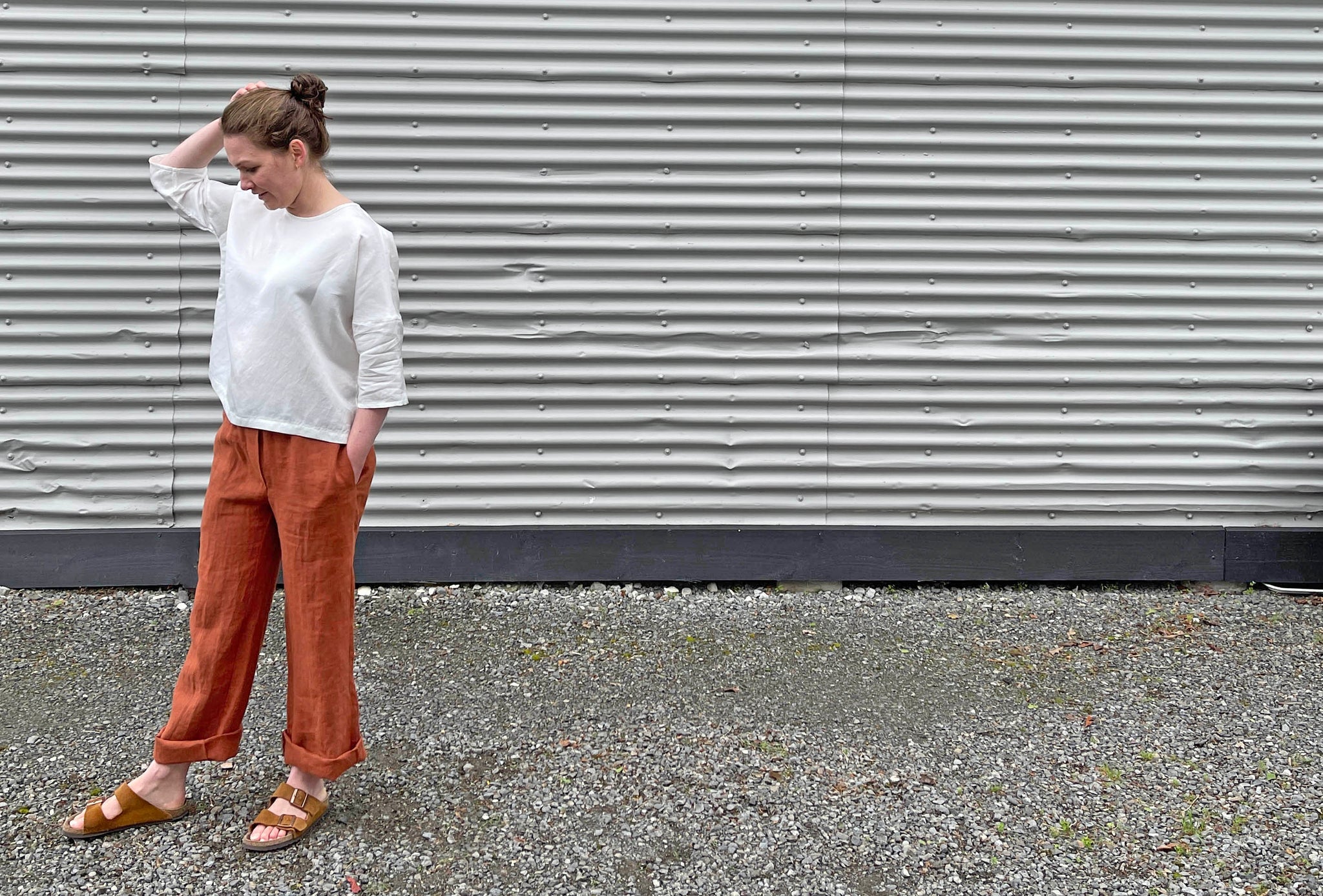 a white lady wearing a white shirt and burnt sienna coloured trousers. They are between orange and brown. It is a side profile of the woman with her hand in her hair.