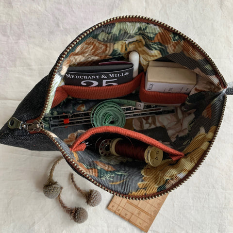a picture of an open sewing-ouch. It has a wide open mouth and is taken from above looking down into it. The pouch is filled with boxes of pins, a tape measure and thread. The pouch is  on an ivory linen background