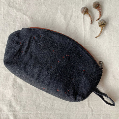 a shot showing the exterior of the pouch in its entirety. it is a dark denim blue with a loop on the right side. It has a gentle curve where the zip is inserted and a flat bottom and is a rectangular shape.